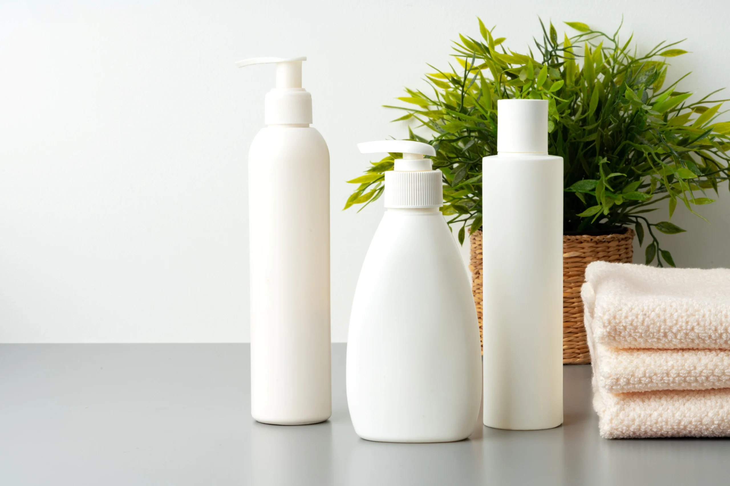 Minimalist white cosmetic bottles placed beside towels and green plant.