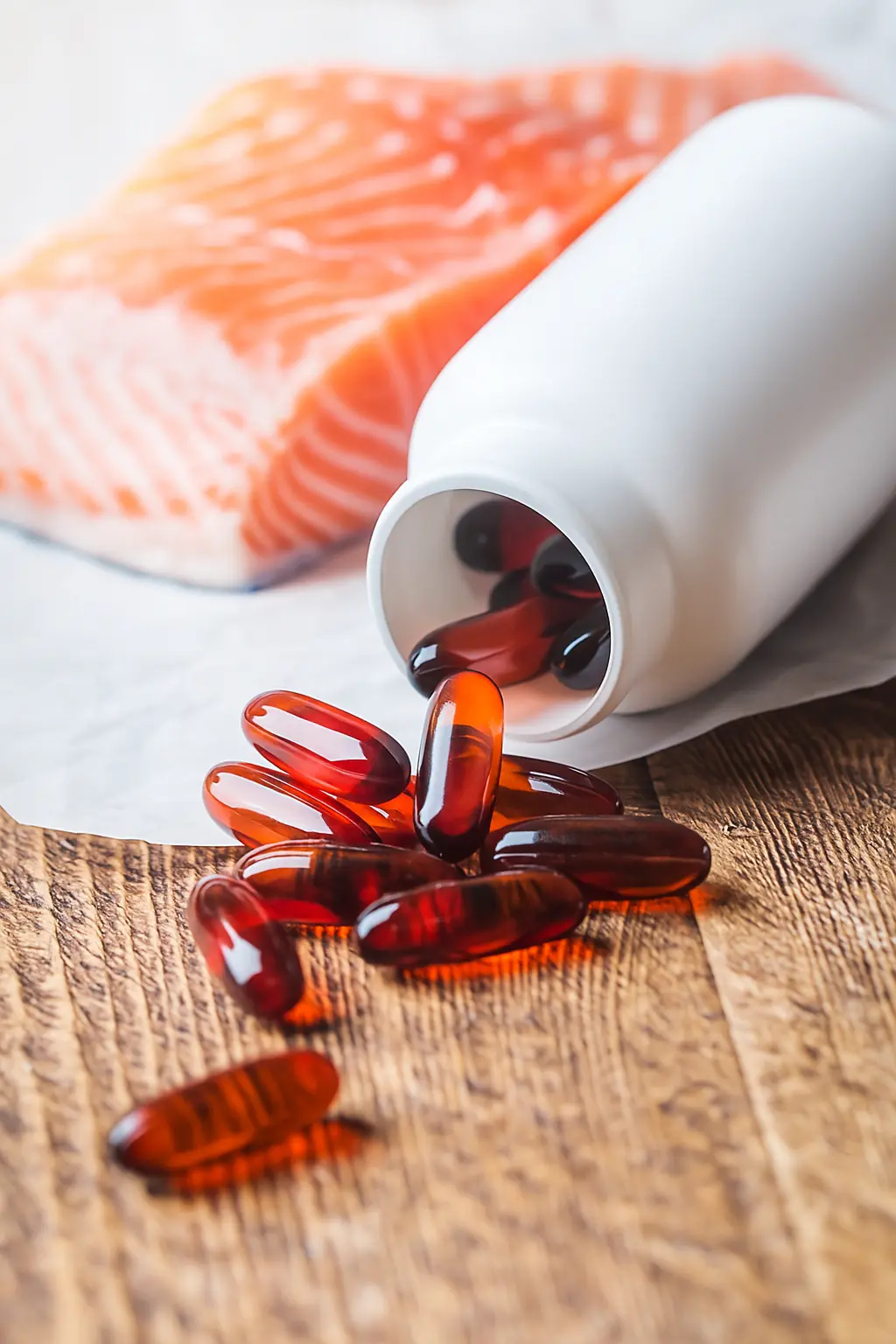 Fish oil capsules spilling from a white bottle beside a raw salmon fillet, showing natural sources of marine oils.