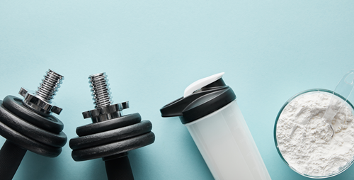 Dumbbells, a white shaker bottle, and a bowl of protein powder with a scoop on a light blue background.