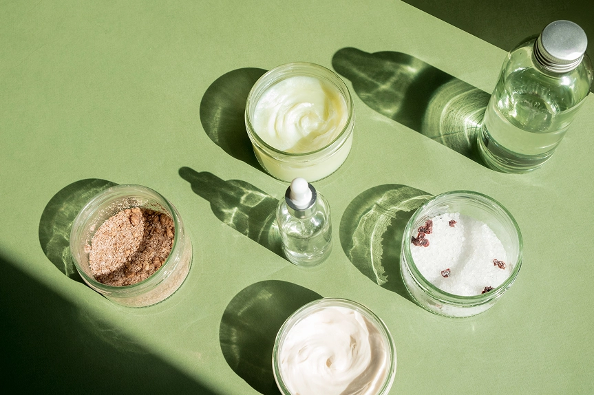 Flatlay of skincare formulation ingredients — cream jars, dropper serum bottle, powder and crystal textures on green background